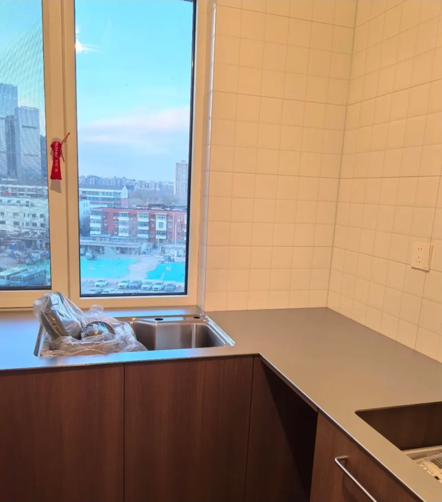 Integrated stainless steel sink welded into a 5mm solid steel countertop, positioned in front of a large window with a city view.
